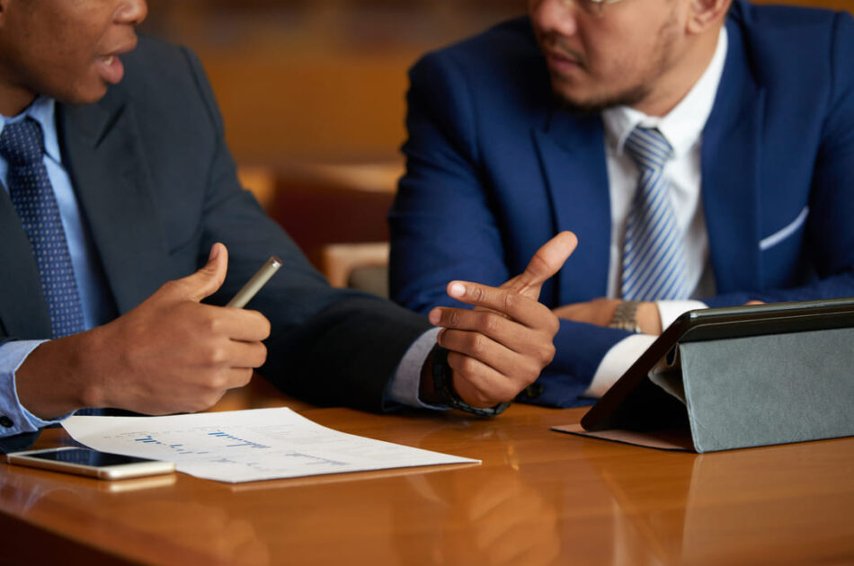 Dois homens com roupas sociais estão conversando em uma mesa. Em cima dela, tem um tablet com a tela na diagonal.