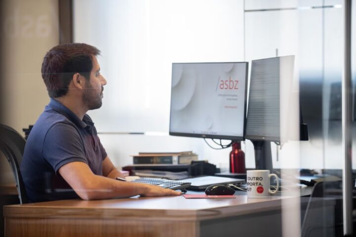 Direito empresarial: homem em sua mesa de escritório mexendo no computador. Na tela, vemos o logo do /asbz.